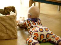 Baby ligger på maven på gulvet. Baby har en hvid, orange og lilla dragt på. Baby leger med en bamse.