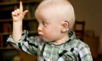 En dreng på 2 år med blå høreapparater sidder med en en hævet pegefinger og siger noget.